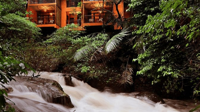 A view of cottages nestled in greenery, from the rocks near the water stream | Vythiri Resort, Wayanad